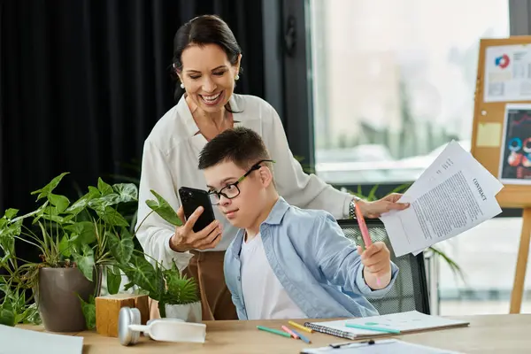 A mother works at her desk in an office while her son, who has Down Syndrome, uses a smartphone. — Stock Photo