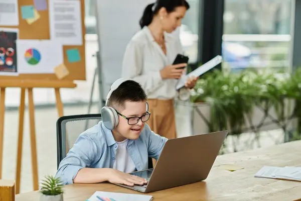 Un niño con síndrome de Down trabaja en una computadora portátil en una oficina en casa mientras su madre trabaja cerca. - foto de stock