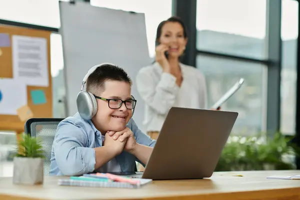 Un ragazzo con la sindrome di Down si siede su una scrivania con un computer portatile mentre sua madre lavora nelle vicinanze. — Foto stock
