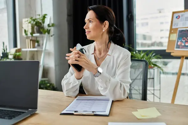 Una mujer con una camisa blanca se sienta en un escritorio en una oficina, sosteniendo un teléfono, mirando pensativo. - foto de stock