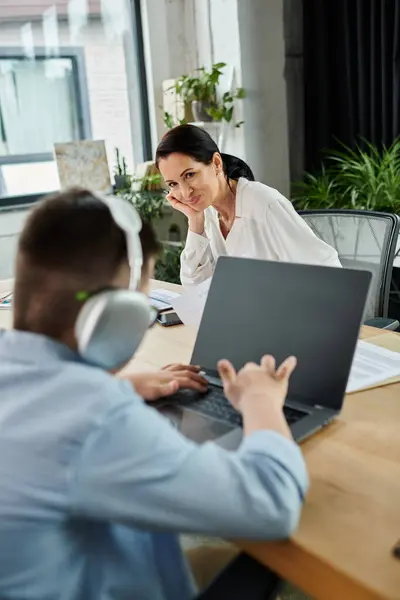Une mère regarde son fils, qui a le syndrome de Down, travailler sur un ordinateur portable dans leur bureau. — Photo de stock