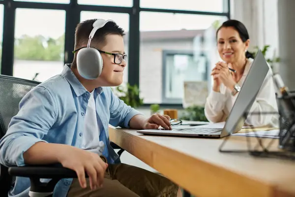 A young boy with Down syndrome sits at a desk in an office, wearing headphones and using a laptop, while his mother works nearby. — Stock Photo