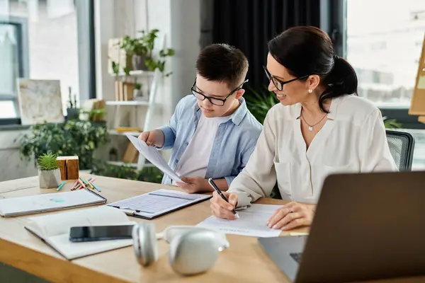 A mother, working on her laptop while her son with Down syndrome does schoolwork, both wearing glasses, showing inclusivity at work. — Stock Photo