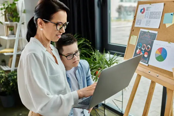 A mother works on her laptop while her son with Down syndrome looks on, showcasing a supportive and inclusive environment. — Stock Photo
