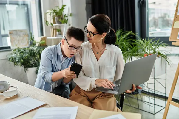 A mother works on her laptop while her son with Down syndrome looks at his phone. — Stock Photo