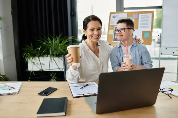 A mother works on her laptop while her son, who has Down syndrome, enjoys a milkshake next to her. — Stock Photo