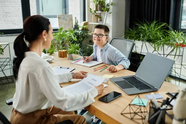 A mother works at her desk while her son with Down syndrome colors at the table. — Stock Photo