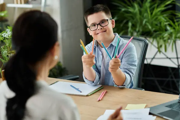 A mother works at her desk while her son with Down syndrome sits beside her, holding up colorful pencils. — Stock Photo