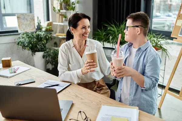 A mother works at her desk in an office while her son with Down syndrome stands next to her, both enjoying drinks. — Stock Photo