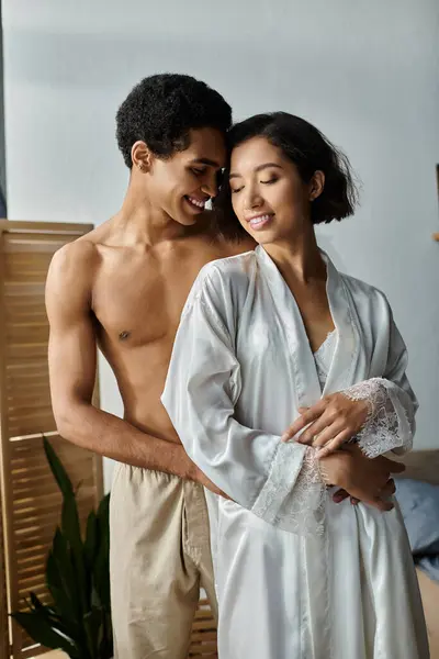 An Asian woman in white robe embraces an African American man in a bedroom, close with faces inches apart, showing intimacy. — Stock Photo