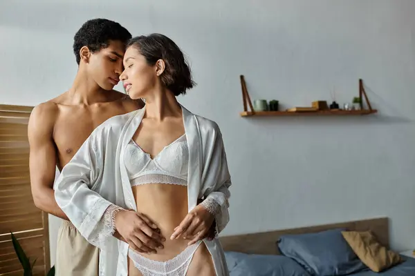 Una joven asiática vestida con una túnica blanca y un hombre afroamericano abrazan y se besan en un dormitorio. - foto de stock