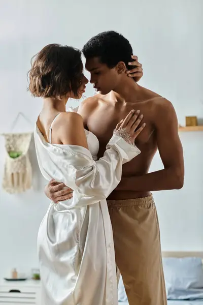 A young Asian woman and an African American man share a loving embrace in a bedroom setting. — Stock Photo