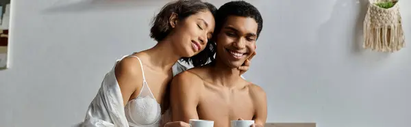 A young multicultural couple enjoys a cozy morning together in their bedroom, sharing a cup of coffee and a loving embrace. — Stock Photo