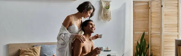 Una giovane coppia multiculturale trascorre del tempo insieme in una camera da letto. La donna è seduta su un letto mentre l'uomo poggia la testa sulla sua spalla. — Foto stock