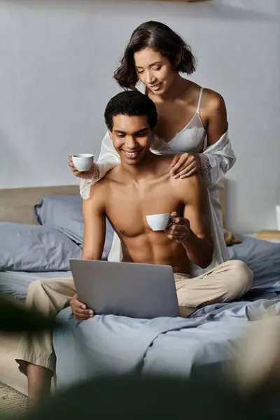 A young interracial couple enjoys a relaxing morning together in their bedroom, sipping coffee and browsing a laptop. — Stock Photo