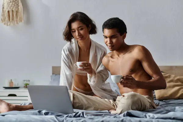 A young, multicultural couple enjoy their morning coffee while sitting on a bed and looking at a laptop. — Stock Photo