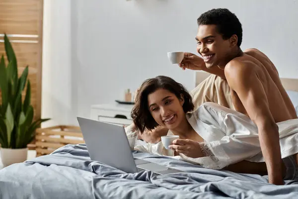 Um jovem casal multicultural gosta de café e um laptop juntos na cama. — Fotografia de Stock