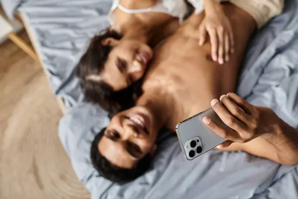 A young multicultural couple relaxes in bed, taking a selfie with a smartphone. — Stock Photo