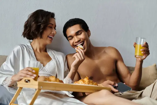 Ein junges multikulturelles Paar frühstückt im Bett, lacht und teilt einen leichten Moment. — Stockfoto