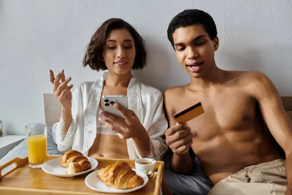 A young multicultural couple enjoys a relaxed morning in bed, sharing breakfast and browsing on their phone. — Stock Photo