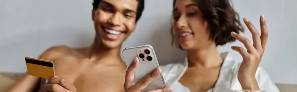 A young, multicultural couple shares a laugh in their bedroom, the man holding a credit card and the woman a phone. — Stock Photo
