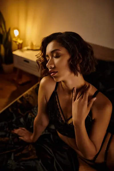 Un'immagine sensuale di una giovane asiatica in lingerie nera, seduta su un letto e che guarda giù con un'espressione seducente. — Foto stock
