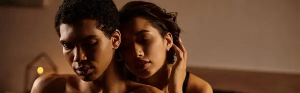 A young Asian woman in black lingerie seduces an African-American man in bed, their faces illuminated by soft, warm light. — Stock Photo