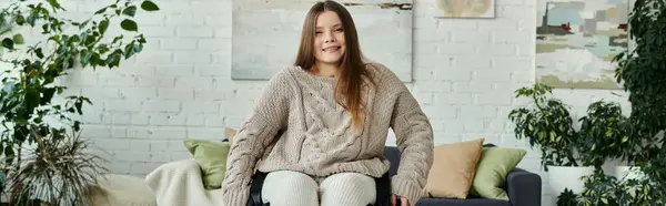 Eine junge Frau mit langen Haaren sitzt in einem Strickpullover in ihrem Haus im Rollstuhl und lächelt in die Kamera. — Stockfoto