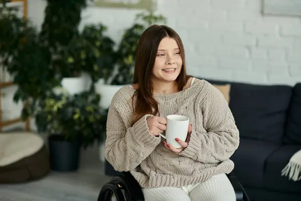 Una giovane donna sulla sedia a rotelle si siede a casa, sorridendo e tenendo in mano una tazza di caffè. — Foto stock