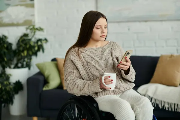 Eine junge Frau sitzt zu Hause im Rollstuhl und schaut auf ihr Smartphone, während sie einen Becher hält. — Stockfoto