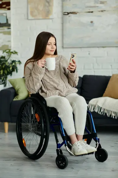 Eine junge Frau im Rollstuhl sitzt zu Hause, genießt eine Tasse Kaffee und schaut auf ihr Handy.. — Stockfoto