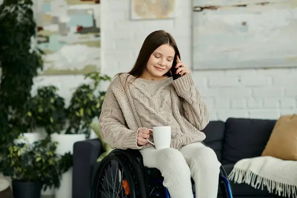 Una giovane donna su una sedia a rotelle si siede a casa in un confortevole soggiorno, tenendo in mano una tazza e parlando al telefono. — Foto stock