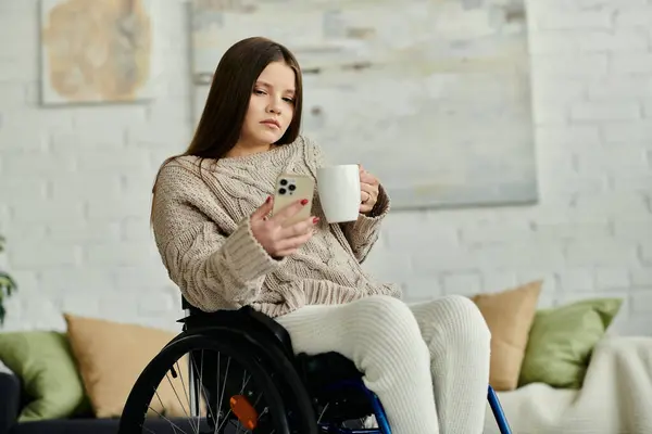 Молода жінка в інвалідному візку зручно сидить вдома, насолоджуючись чашкою чаю і прокручуючи свій телефон. — стокове фото