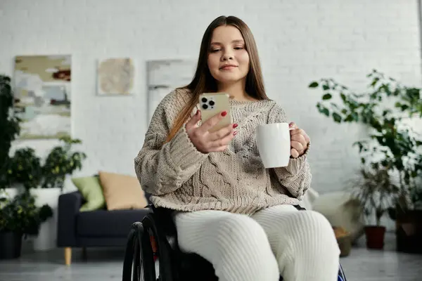 A woman in a wheelchair uses her phone while sitting in her living room, enjoying a cup of coffee. — Stock Photo