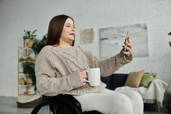 A young woman in a wheelchair smiles as she takes a selfie while enjoying a morning cup of coffee in her living room. — Stock Photo