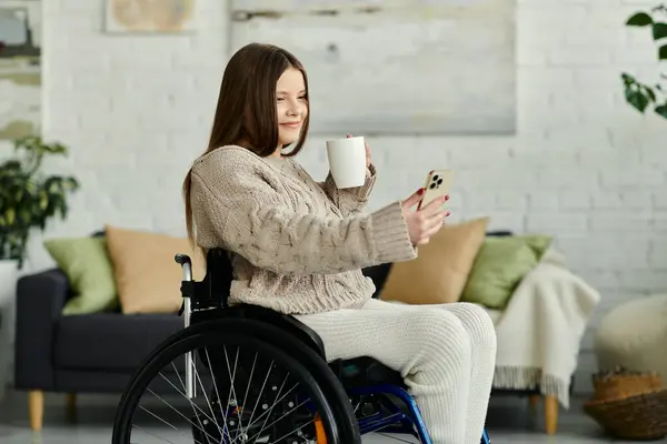 A young woman in a wheelchair sits at home, enjoying a cup of coffee and browsing her phone. — Stock Photo