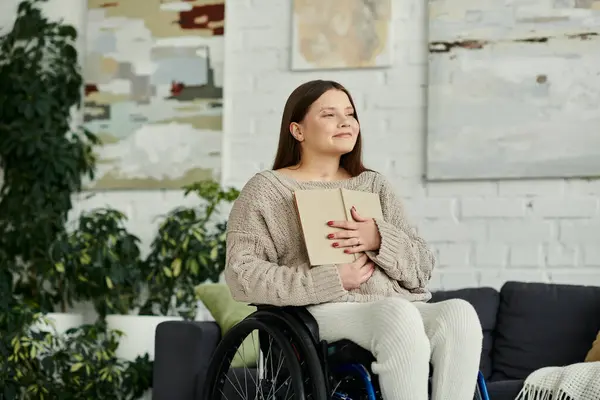 Eine junge Frau sitzt im Rollstuhl in ihrem Wohnzimmer und hält ein Buch vor der Brust. — Stockfoto