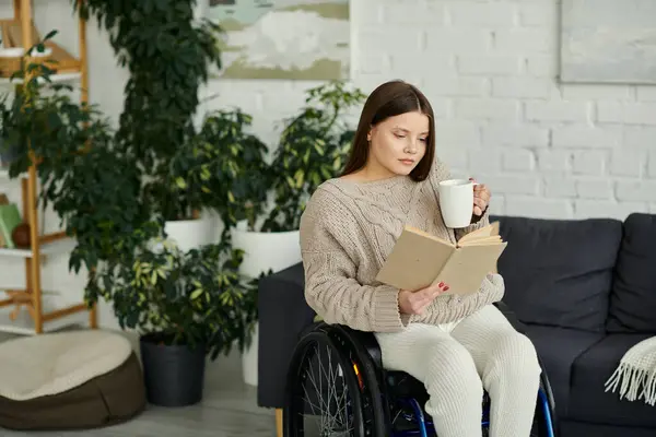 Eine junge Frau im Rollstuhl sitzt in ihrem Wohnzimmer und genießt eine Tasse Kaffee während sie ein Buch liest. — Stockfoto