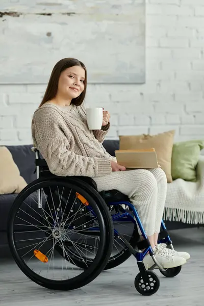 Una giovane donna sulla sedia a rotelle si siede a casa, gustando una tazza di caffè e un libro. — Foto stock