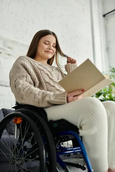 Eine junge Frau im Rollstuhl sitzt zu Hause, liest ein Buch und lächelt. — Stockfoto