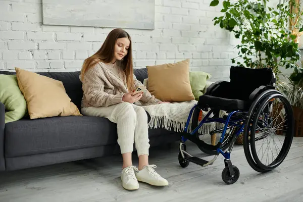 A young woman in a wheelchair sits on a couch at home, looking at her phone. — Stock Photo