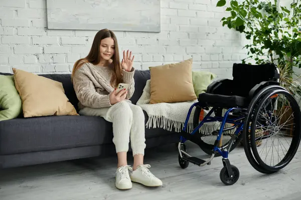 A young woman sits on a couch at home, using her smartphone while her wheelchair is nearby. — Stock Photo