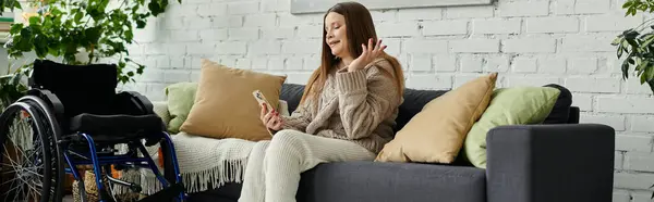 Eine junge Frau sitzt auf einer Couch in ihrem Haus und benutzt ihr Handy. Ihr Rollstuhl ist im Hintergrund. — Stockfoto
