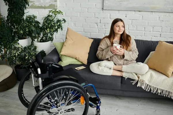 Eine junge Frau im Rollstuhl sitzt auf einer Couch, hält eine Tasse Kaffee in der Hand und schaut aus dem Fenster. — Stockfoto