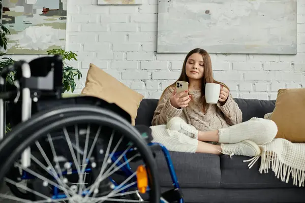 Eine junge Frau im Rollstuhl entspannt auf ihrem Sofa, genießt eine Tasse Kaffee und surft auf ihrem Handy.. — Stockfoto