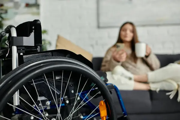 A close-up of a wheelchairs wheel, with a young woman relaxing at home in the background. — Stock Photo