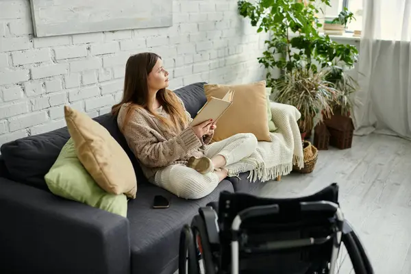 A young woman in a wheelchair sits on a couch, engrossed in a book, enjoying a quiet moment at home. — Stock Photo