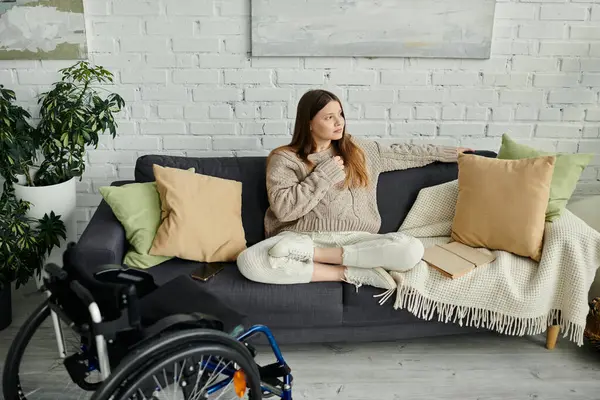 A young woman sits in a wheelchair, relaxed on a couch in her home. — Stock Photo