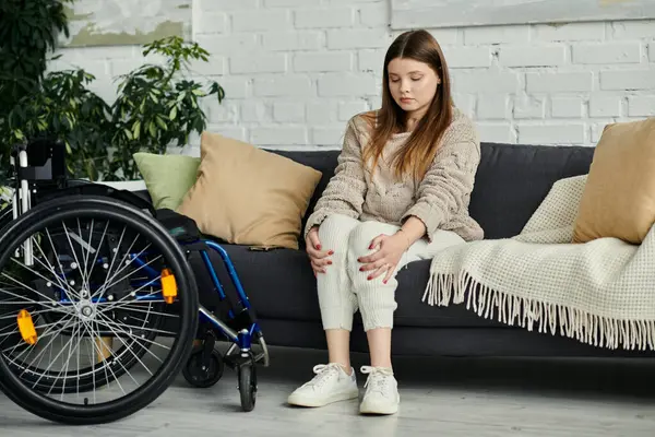 A young woman in a wheelchair sits on a couch at home, resting and looking thoughtful. — Stock Photo