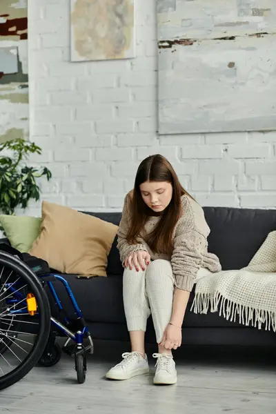 A young woman in a wheelchair puts on her shoes while sitting on a couch at home. — Stock Photo